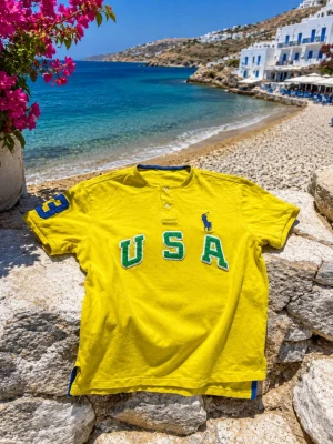 Gul t-shirt från Ralph Lauren - Gul t-shirt från Ralph Lauren med stora bokstäver 'USA' i grönt och vitt framtill. Blå logga på bröstet och blå detalj på ärmen. Kortärmad modell med knappar vid halsen. Perfekt för en sportig look.