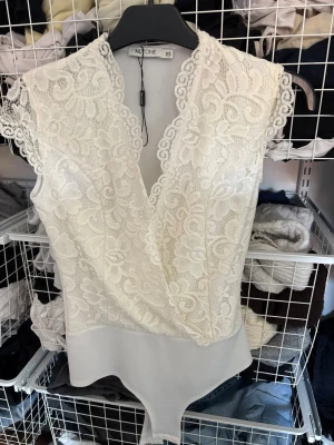 Vit spetsbody från NLY One XS - Säljer en superfin vit body från NLY One i storlek XS. Framsidan har vacker spets med blommönster och omlott-ringning, medan baksidan är slät och enkel. Korta spetsärmar ger en romantisk touch. Perfekt att styla till jeans eller kjol!