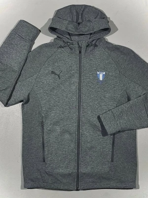 Grå hoodie från Puma Malmö FF - Snygg grå hoodie från Puma med Malmö FF-logga på bröstet och dragkedja framtill. Tröjan har två fickor med dragkedja, huva och sportig design. Perfekt för dig som vill visa vilket lag du hejar på lite stilrent och diskret. Materialet är mjukt och stretchigt för extra komfort samt Pumas ”DRYCELL”.