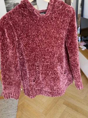Vinröd chenille hoodie från ONLY - Mysig vinröd hoodie från ONLY i supermjuk chenille. Tröjan har huva, långa ärmar och en stor magficka framtill. Perfekt för dig som gillar en avslappnad och trendig look. Materialet är glansigt och lent, vilket gör den extra skön att ha på sig.