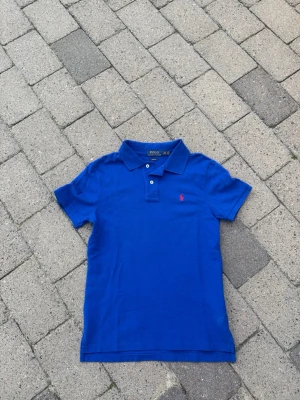 Blå pikétröja från Polo Ralph Lauren - Klassisk blå pikétröja från Polo Ralph Lauren med röd broderad logga på bröstet. Tröjan har korta ärmar, krage och knappar framtill. Slim fit-modell i bomull som ger en snygg och bekväm passform.