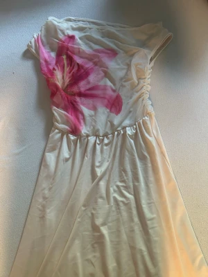 Vit klänning med rosa blomma - Elegant vit klänning med en stor rosa blommig detalj på bröstet. Klänningen har en asymmetrisk axel och är tillverkad i ett glansigt material som ger en lyxig känsla. Perfekt för sommarens tillfällen.