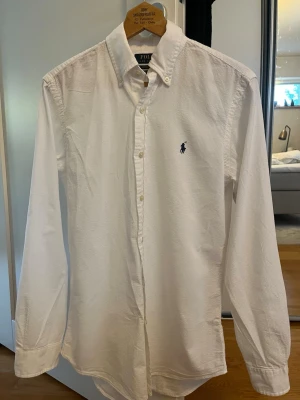 Vit skjorta från Polo Ralph Lauren - Klassisk vit skjorta från Polo Ralph Lauren med broderad logga på bröstet. Skjortan har lång ärm, knappar framtill och är i bomull. Custom fit-modell som sitter snyggt och passar till många olika stilar.