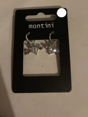 Silverörhängen med hjärtformad sten - Snygga örhängen från Montini i silverfärg med hjärtformad klar sten och pil-detalj. Perfekta för att ge din look lite extra edge och glans. Metall och glittrande sten ger en cool och trendig vibe.