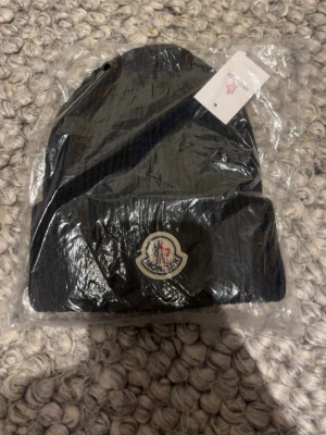 Svart Moncler mössa med logga - Snygg svart ribbstickad mössa från Moncler med klassisk uppvikt kant och broderad logotyp framtill. Perfekt accessoar för att hålla värmen och ge din outfit en exklusiv touch. Mössan är i mjukt material och har en stilren design.
