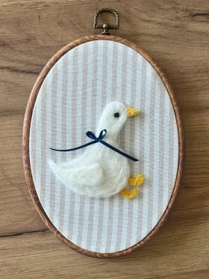 Broderad tavla med filtad anka - Handgjord oval tavla med en söt vit anka i ull, dekorerad med en mörkblå rosett runt halsen. Bakgrunden är ljus med smala beige ränder och ramen är i trä. Perfekt som gullig inredningsdetalj till rummet.