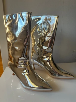 Silvriga metallic stövlar med klack - Säljer ett par nya & oanvända höga stövlar i metallic-silver med spetsig tå och smal klack. Klacken är medelhög dvs mellan 5-6,5 cm.. Stövlarna har en glansig finish och dragkedja på insidan. Perfekta för dig som vill sticka ut och ge outfiten en vrålball touch. Strl 41. Kvalité:Polyuretan. Kommer i sin original-kartong. Aldrig ens provade, bara legat i sin medföljande låda och i silkespapper. 
