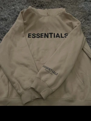 Beige Essentials hoodie Fear of God - Snygg beige hoodie från Fear of God Essentials med svart tryck på ryggen och ärmen. Hoodien har en klassisk huva och känguruficka framtill. Perfekt för en avslappnad streetwear-look och passar till det mesta i garderoben.