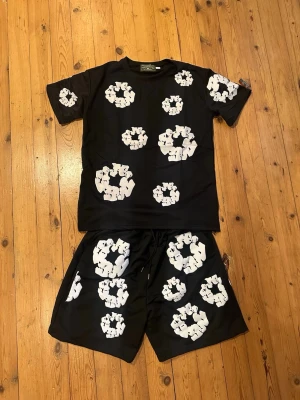 Svart Denim Tears set - Säljer ett svart set från Denim Tears med matchande t-shirt och shorts. Båda plaggen har vita blommönster tryckta över hela ytan. Setet har en avslappnad passform och är gjort i mjukt material, perfekt för en cool och bekväm stil.