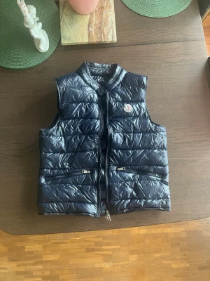 Mörkblå dunväst från Moncler - Snygg mörkblå dunväst från Moncler med glansig finish och klassisk logotyp på bröstet. Västen har två dragkedjefickor framtill och stängs med dragkedja. Perfekt för lager-på-lager och ger en sportig vibe.  