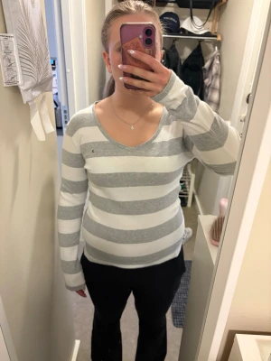 Randig tröja från Tommy Hilfiger  - Snygg långärmad tröja från Tommy Hilfiger med breda grå och vita ränder. Tröjan har v-ringning och en liten broderad logga på bröstet. Perfekt för en avslappnad och stilren look.