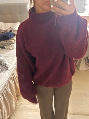 Mysig vinröd stickad polotröja - Vinröd stickad tröja med hög krage och oversized passform. Tröjan har långa ärmar och är gjord i ett mjukt, fluffigt material som känns riktigt skönt mot huden. Perfekt för kalla dagar när du vill vara både varm och snygg.