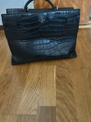 Svart portfölj Guess croco-mönster - Säljer en svart portfölj från Guess med croco-mönstrad yta och stilrena handtag. Väskan har metallfärgade detaljer och ett elegant, strukturerat utseende. Perfekt för dig som vill ha en snygg och trendig portfölj till skolan eller jobbet.