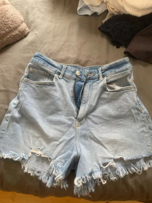 Ljusblå högmidjade shorts - Säljer ett par ljusblå denimshorts med hög midja och fransig, rå kant nertill. Klassisk femficksmodell med knapp och dragkedja framtill. Perfekta för sommaren och ger en avslappnad vibe.