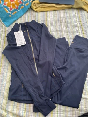 Mörkblått träningsset från Lululemon - Snyggt mörkblått träningsset från Lululemon med matchande zip-jacka och tights. Jackan har dragkedja och fickor med dragkedja, tightsen har Lululemon-logga på höften. Materialet är stretchigt och följsamt, perfekt för träning eller yoga.