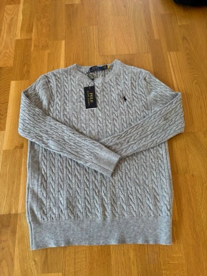 Grå kabelstickad tröja Polo Ralph Lauren - Snygg grå kabelstickad tröja från Polo Ralph Lauren med rund halsringning och klassisk broderad logga på bröstet. Tröjan har ribbade muddar och är tillverkad i mjukt material som passar perfekt till lager-på-lager-stil.