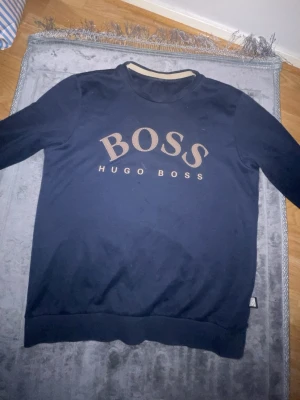 Svart Hugo Boss t-shirt med logga - Snygg svart t-shirt från Hugo Boss med stor beige logga på bröstet. Klassisk rund halsringning och rak passform. Perfekt för dig som gillar stilrena och trendiga plagg med tydlig branding.