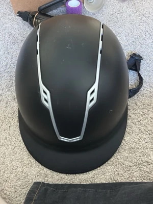 Svart ridhjälm från John Whitaker - Snygg svart ridhjälm från John Whitaker med matt finish och silverfärgade ventilationsdetaljer. Hjälmen har justerbart hakband och modern design med logga på sidan. Perfekt för dig som vill ha både stil och säkerhet i stallet.