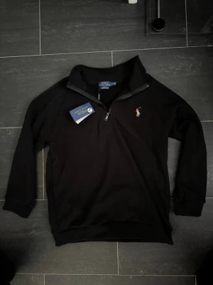 Svart half-zip tröja från Polo Ralph Lauren - Svart långärmad tröja från Polo Ralph Lauren med klassisk half-zip och broderad logga på bröstet. Tröjan har ribbade muddar och en mjuk bomullskänsla. Perfekt för en stilren och avslappnad look. Passar för L och M 