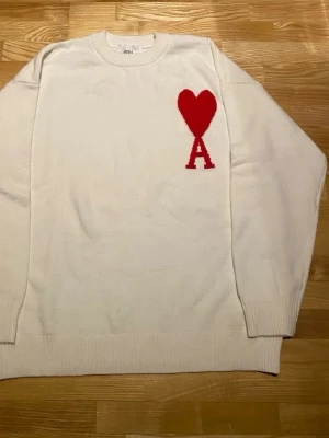 Vit stickad tröja från Ami Paris - Vit stickad tröja från Ami Paris med rött hjärta och bokstaven A broderat på bröstet. Tröjan har rund halsringning, långa ärmar och ribbade muddar. Tillverkad i mjuk ullblandning som känns lyxig och skön.