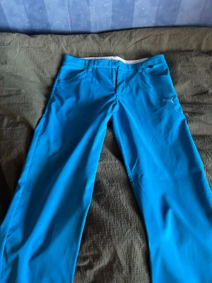 Blå golf byxor från Puma - Snygga blå golf byxor från Puma med raka ben och klassisk passform. Byxorna har framfickor, bakfickor och Puma-logga på framsidan. Perfekta för golfspelare som vill sticka ut lite extra på bannan. Materialet känns mjukt och rörligt. Det följer med en extra knapp!