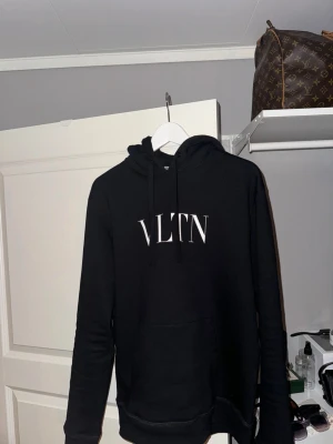 Svart hoodie från Valentino VLTN - Snygg svart hoodie från Valentino med VLTN-tryck i vitt på bröstet. Klassisk modell med huva och dragsnören samt en stor magficka. Tillverkad i mjukt material som känns skönt mot huden. Perfekt för en trendig och avslappnad stil.