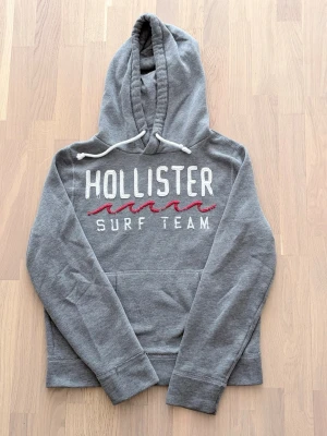 Hollister hoodie - En riktigt fet, stilren, grå hoodie från Hollister med vit och röd brodyr på bröstet där det står 'Hollister Surf Team'.                                                   - Storlek: Medium.                                                        - Paket lämnas alltid in inom 24h efter köp!