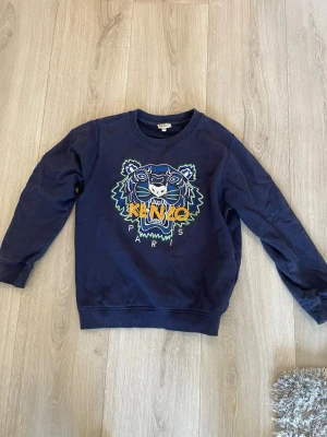 Mörkblå Kenzo stickad tröja tiger - Mörkblå stickad tröja från Kenzo med ikoniskt tigertryck och färgglada detaljer framtill. Rund halsringning och ribbade muddar vid ärmslut och nederkant. Perfekt för dig som gillar streetstyle och vill sticka ut.