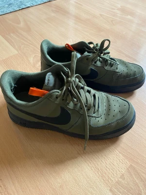 Nike Air Force 1 olivgröna sneakers - Snygga Nike Air Force 1 sneakers i olivgrönt med svart swoosh och mörk sula. Klassisk låg modell med perforerad tå och gröna skosnören.                                                  Innermått 25 cm