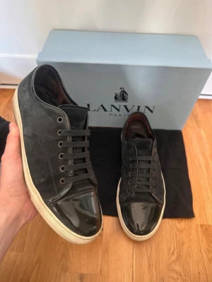 Svarta Lanvin  - Finns en repa på vänstra sko på glans så ni vet så det inte blir missförstånd ifall man köper. Snygga svarta sneakers från Lanvin med ovandel i mocka och lackad tå. Skorna har svarta skosnören och vit sula som ger en clean kontrast. Perfekta för dig som gillar stilrena och exklusiva sneakers med unika materialval.