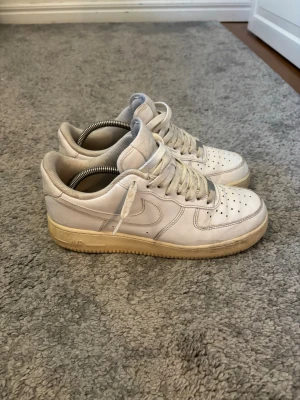 Nike Air Force 1 vita sneakers - Klassiska Nike Air Force 1 sneakers i vitt läder med perforerad tå, vit sula och ikonisk Swoosh på sidan. Skorna har lågt skaft och platt gummisula. Perfekta för en clean och stilren look.