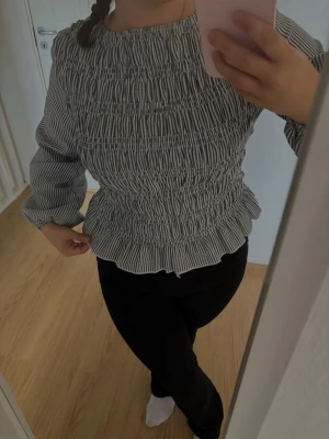Randig smockad blus med långa ärmar - Säljer en snygg blus med smockad midja och rynkade detaljer. Blusen är vit med svarta tunna ränder och har långa ärmar med resår vid ärmslut. Den har en rund halsringning och en figurnära passform i midjan. Perfekt för en trendig look.