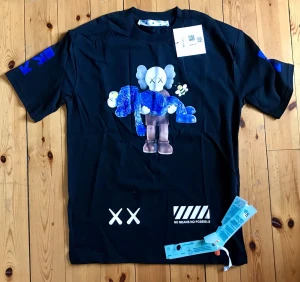 Svart Off-White x KAWS t-shirt - Svart t-shirt från Off-White i samarbete med KAWS. Framsidan har en stor färgglad KAWS-figur och tryck med vita detaljer. Baksidan har Off-Whites ikoniska pilar med KAWS-motiv. T-shirten är kortärmad och i mjuk bomull.