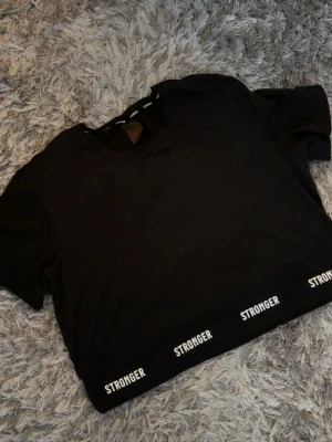 Svart crop top från Stronger - Svart crop top från Stronger med korta ärmar och bred resår nertill där det står 'STRONGER' i vitt flera gånger. Perfekt för träning eller gymmet, tillverkad i stretchigt material som sitter skönt på kroppen.
