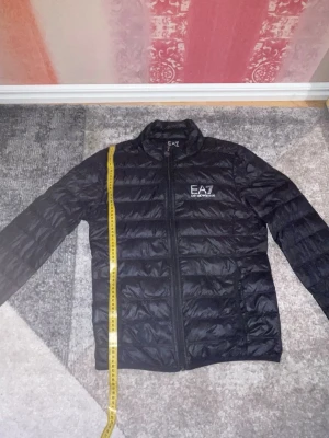 Svart pufferjacka från EA7 Armani - Snygg svart pufferjacka från EA7 Emporio Armani med diskret glansig finish. Jackan har dragkedja framtill, ståkrage och två sidofickor. EA7-logga broderad på bröstet. Perfekt för kalla vinterdagar och riktigt trendig stil.