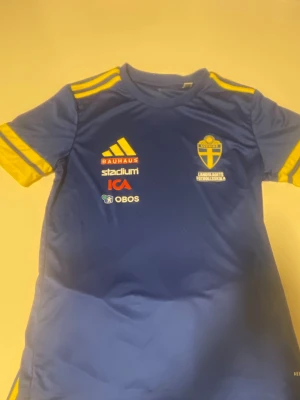 Blå Sverige fotbollströja Adidas M - Säljer en blå fotbollströja från Adidas i storlek M, med gula detaljer och Sveriges landslagsemblem på bröstet. Tröjan har tryckta sponsorer och är i slim fit-modell. Perfekt för dig som vill stötta landslaget på match eller träning.