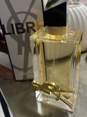 Yves Saint Laurent Libre Eau de Parfum - Snygg parfymflaska från Yves Saint Laurent, Libre Eau de Parfum. Flaskan är rektangulär i genomskinligt glas med gulddetaljer och svart asymmetrisk kork. Den har en lyxig känsla med YSL-logga i metall på framsidan.