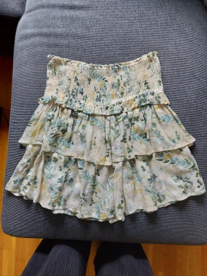 Blommig volangkjol med smock - Supersöt kort kjol med blommigt mönster i grönt, gult och blått på ljusbeige botten. Kjolen har smock i midjan och flera volanger som ger en luftig och somrig känsla. Perfekt för varma dagar och enkel att matcha med en enkel topp.
