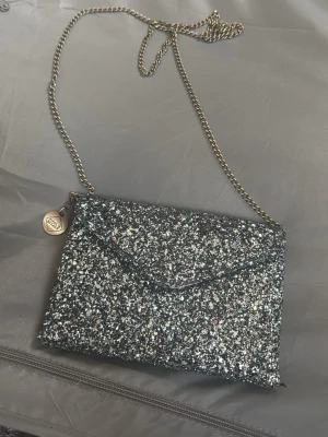 Glittrig silver kuvertväska med kedja - Säljer en blå glittrig kuvertväska från pipol’s bazaar med  kedjerem. Väskan har magnetlås och insida i ljus satin med ett litet innerfack. Perfekt för att addera lite extra bling till din outfit!