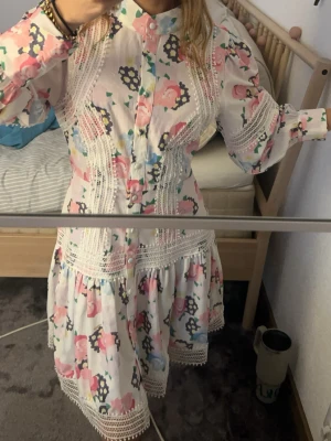 Blommig kortklänning med spetsdetaljer - Superfin kortklänning med blommigt mönster i rosa, blått och gult på vit botten. Klänningen har långa ärmar, knappar framtill och vackra spetsdetaljer längs sömmarna och nederkanten. Perfekt för vår och sommar!