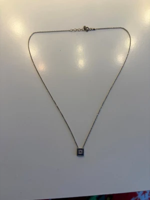 Silverhalsband med fyrkantig berlock - Snyggt halsband i silverfärg med en tunn kedja och en fyrkantig berlock dekorerad med små glittrande stenar. Perfekt för att ge en stilren touch till din outfit. Berlocken har en modern och minimalistisk design.