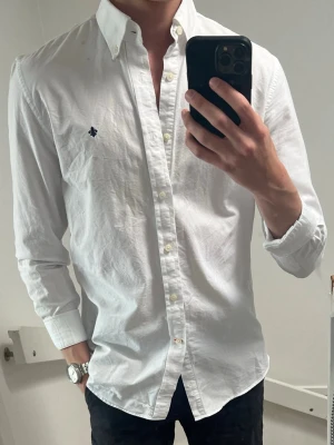 Vit skjorta från Morris - Klassisk vit skjorta från Morris med button-down krage och diskret broderad logga på bröstet. Skjortan har långärmad design, knappar framtill och är tillverkad i bomull. Perfekt för dig som gillar stilrena plagg med snygga detaljer.