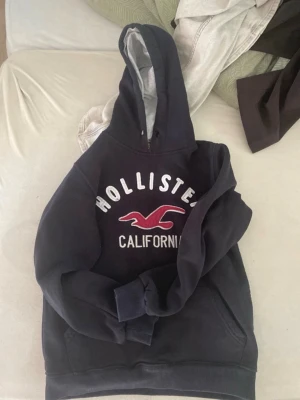 Mörkblå Hollister hoodie med tryck - Svart hoodie från Hollister med vit och röd broderad logga och texten 'Hollister California' på bröstet. Mjuk insida, stor huva och känguruficka framtill. Perfekt för en chill och avslappnad stil.