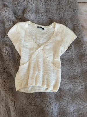 Ljus beige topp med volangkant - Supersöt ljus omlottopp med korta ärmar och volangkant längs v-ringningen. Toppen har en ribbad struktur och är tillverkad i ett mjukt, stretchigt material som känns bekvämt mot huden. knappt använd och storlek xs.💛