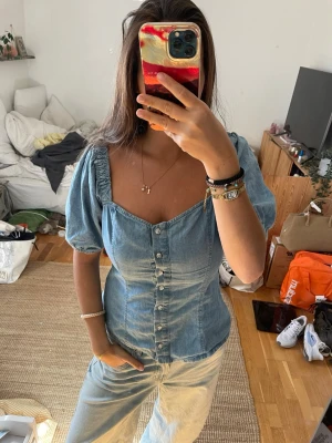 Blå jeansblus med puffärm och knappar - Säljer en snygg blå jeansblus med puffärmar och markerad midja. Toppen har v-ringning, dekorativa knappar framtill och korta ärmar med lätt rynk. Perfekt för dig som gillar en trendig och feminin look.