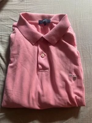 Rosa pikétröja från Gant - Klassisk rosa pikétröja från Gant med krage och korta ärmar. Tröjan har knappslå framtill och en liten broderad logga på bröstet. Perfekt för en avslappnad och stilren look.