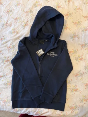 Mörkblå hoodie från Peak Performance - Snygg mörkblå hoodie med dragkedja från Peak Performance. Modellen har huva, två fickor framtill och vit broderad logga på bröstet. Perfekt för en avslappnad och sportig stil. Materialet känns mjukt och bekvämt mot huden.