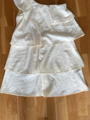 Vit omlottklänning med volanger - Snygg vit kortklänning i omlottmodell med flera lager volanger och oneshoulder-design. Tillverkad i ett glansigt material som ger en lyxig känsla. Perfekt för dig som vill sticka ut med en unik och trendig look.