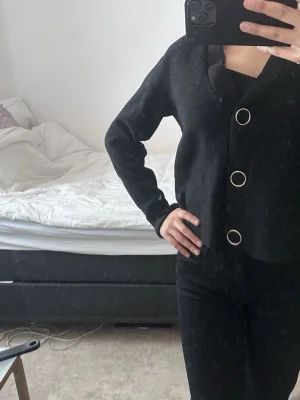 Svart kofta med stora knappar - Stilren svart kofta med tre stora, runda metallknappar framtill. Koftan har v-ringning och långa ärmar med ribbade muddar. Perfekt att slänga över en topp för en enkel men snygg look.