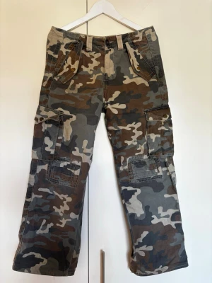 Camo cargobyxor med vida ben - Säljer ett par cargobyxor i klassiskt camomönster i grönt, brunt och beige. Byxorna har flera stora fickor på sidorna och bak, samt vida ben och förstärkta sömmar. En tryckknapp trasig och slitna nedtill men påverkar inte helhetsintrycket! Storlek 29